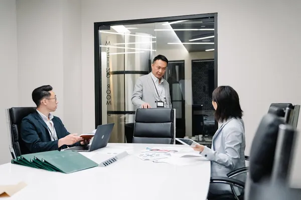 Three Singapore office colleagues mid-discussion in a glass-walled meeting room — one standing and speaking, two seated and listening, with papers and a laptop on a white conference table