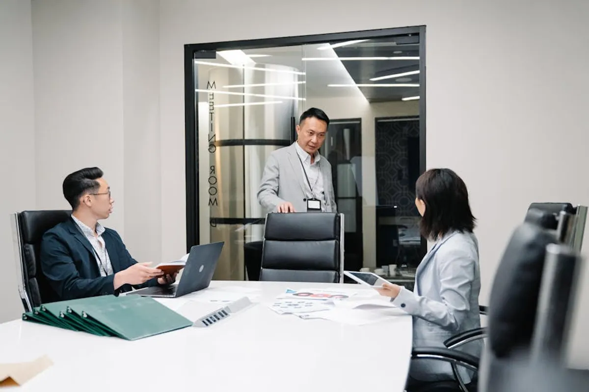 Three Singapore office colleagues mid-discussion in a glass-walled meeting room — one standing and speaking, two seated and listening, with papers and a laptop on a white conference table