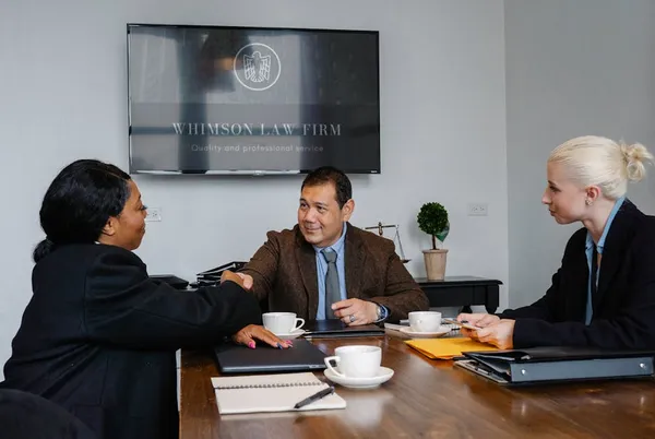 A Singapore account manager and a client mid-conversation across a clean meeting table — listening posture, open laptop with notes, calm professional setting