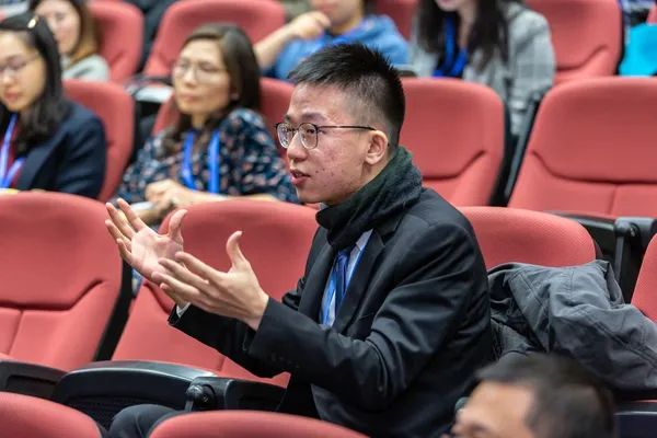 A Singapore presenter standing calmly at the front of a meeting room taking a question from the audience, hands open and listening intently