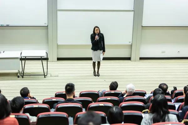 A Singapore presenter on a clean stage with one large headline visible behind them, single hand-held microphone, audience in soft focus — keynote-style setup
